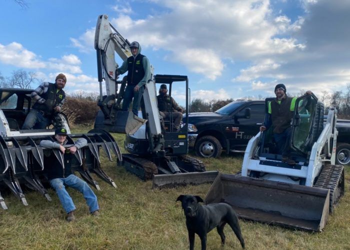 A group of men are standing in a field with tractors and a dog.