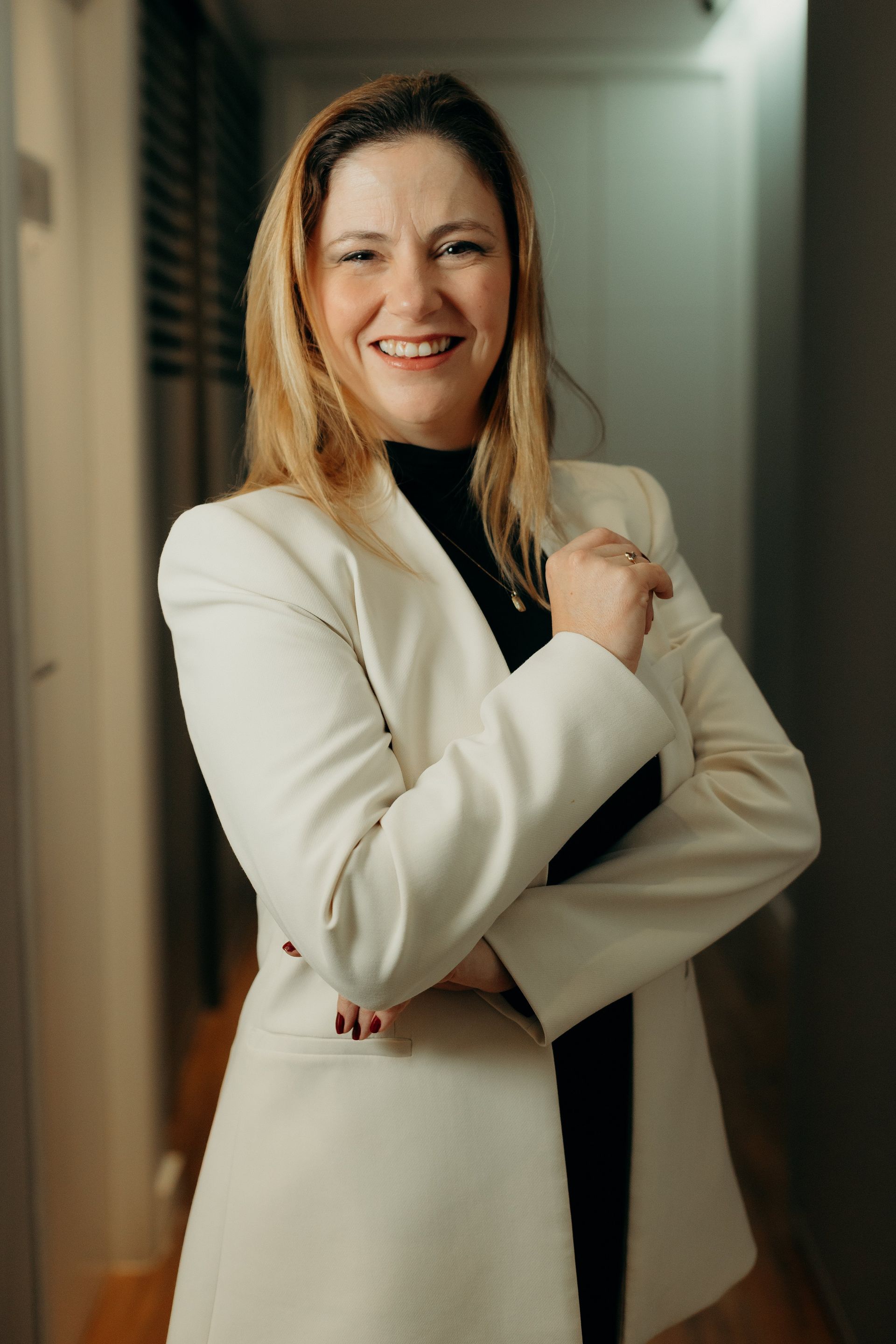 Uma mulher de jaqueta branca está parada em um corredor com os braços cruzados e sorrindo.