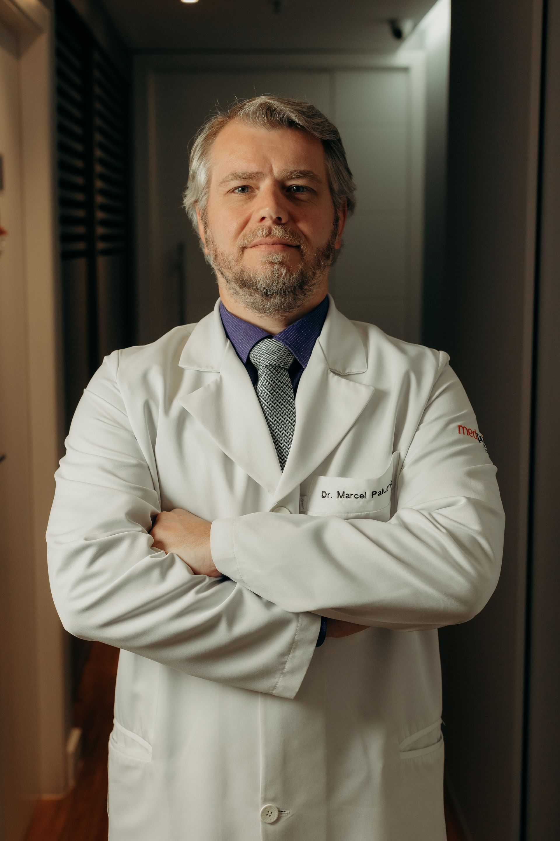 Um homem de jaleco e gravata está de pé com os braços cruzados.