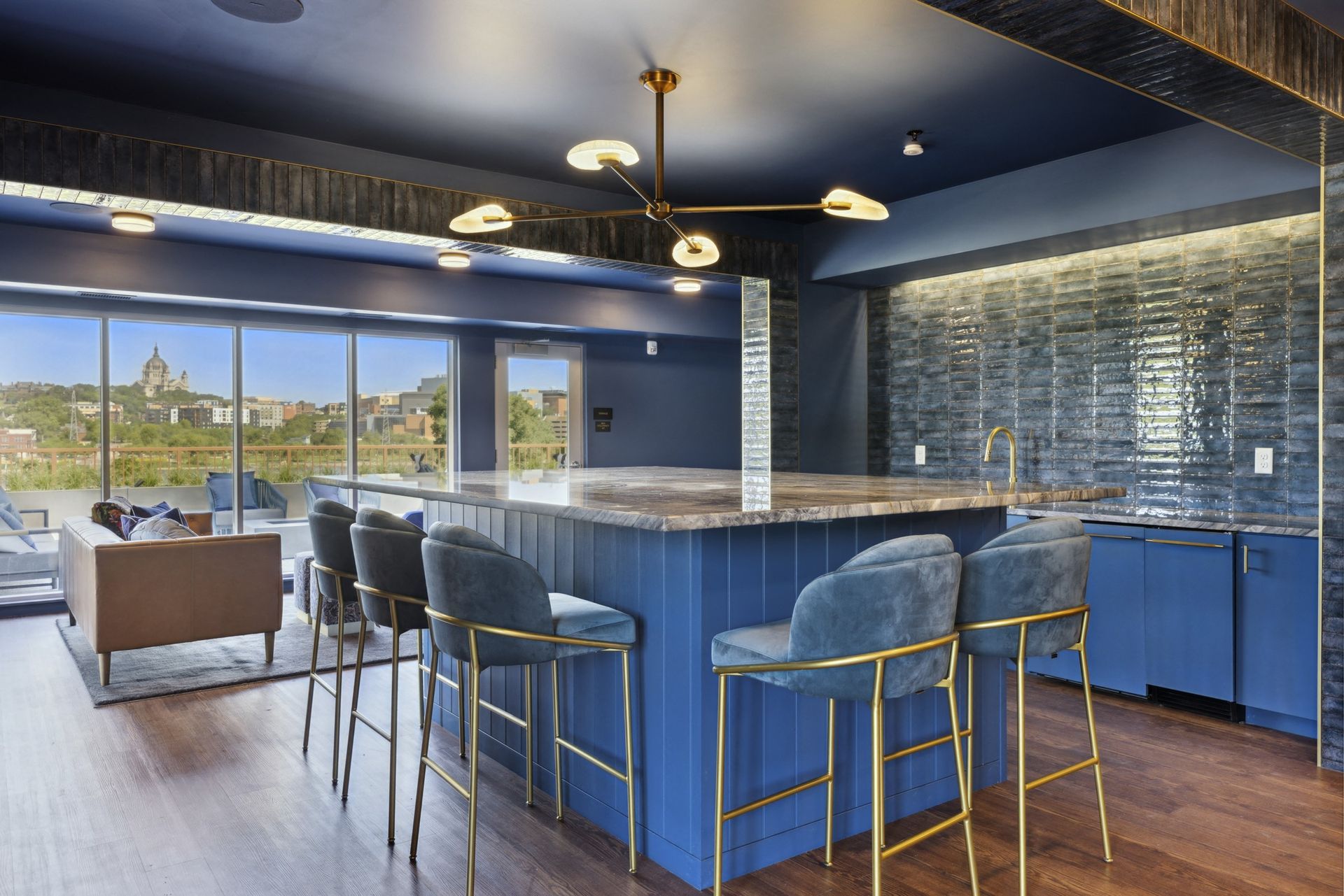 Interior of a modern communal lounge with a blue island bar, gold stools, and large city-view windows at Esox House in St. Paul, MN.