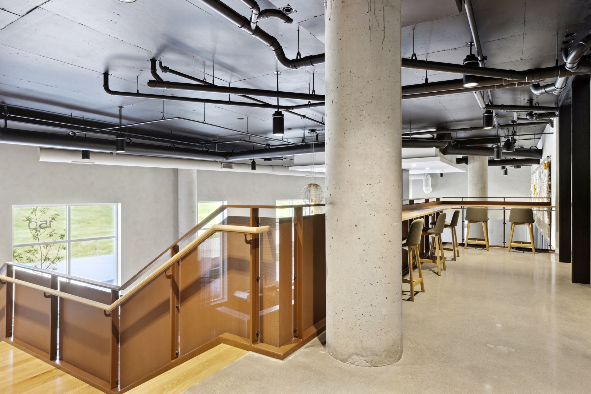 Open indoor communal lounge with a staircase, a tall concrete pillar, and exposed ceiling pipes at Esox House in St. Paul, MN.