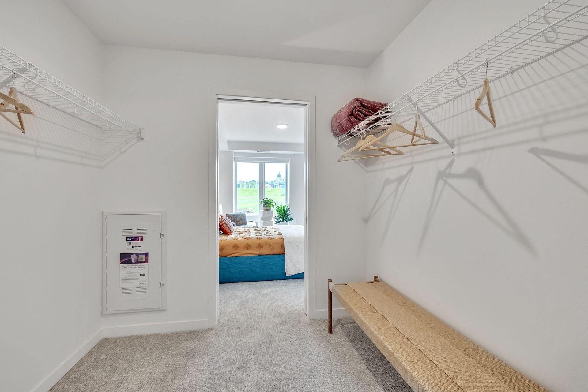 Walk-in closet with white wire shelving on both sides, a beige bench, and a doorway to a bedroom at Esox House in St. Paul, MN.