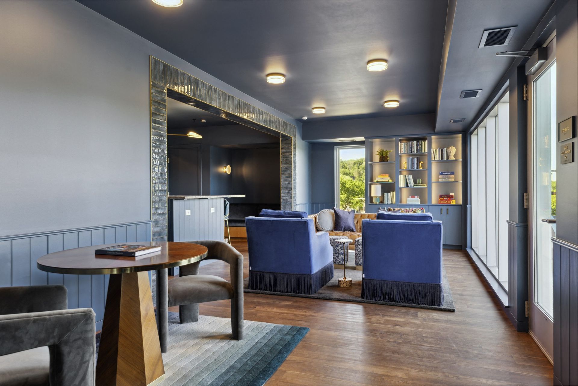 Blue-toned communal lounge with sofas, a bookshelf wall, and large windows at Esox House in St. Paul, MN.