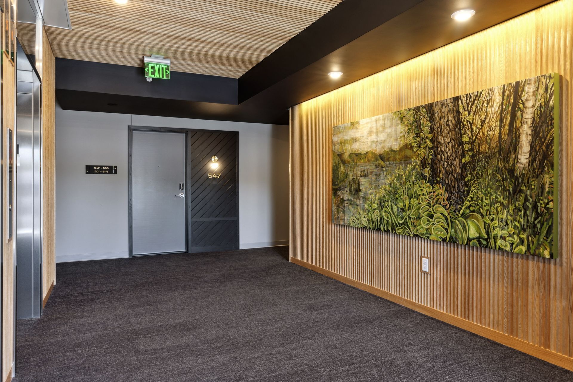 Interior apartment community hallway with wood slat walls, a large landscape painting, carpet, and an exit sign at Esox House in St. Paul, MN.