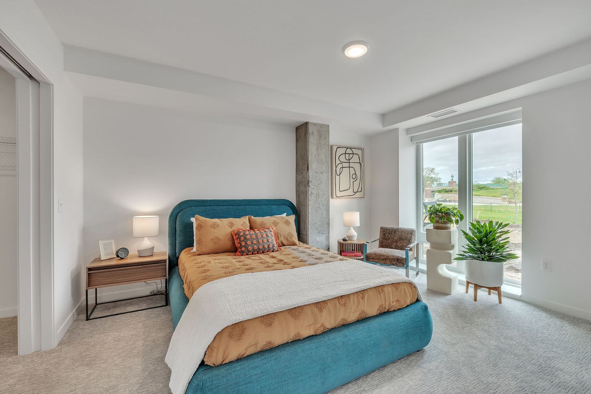 Bright bedroom with teal upholstered bed, nightstands, and plants by a large window at Esox House in St. Paul, MN.