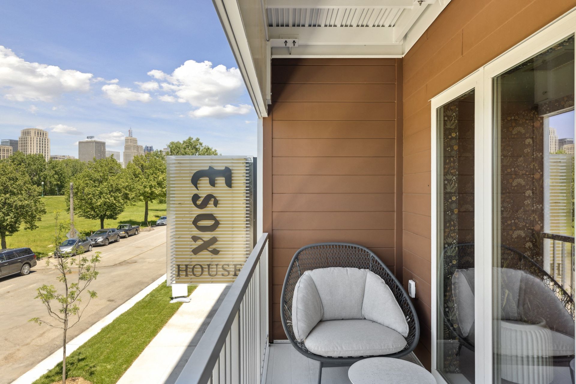 Balcony with a round gray chair and glass door on brown siding, overlooking city skyline at Esox House in St. Paul, MN.