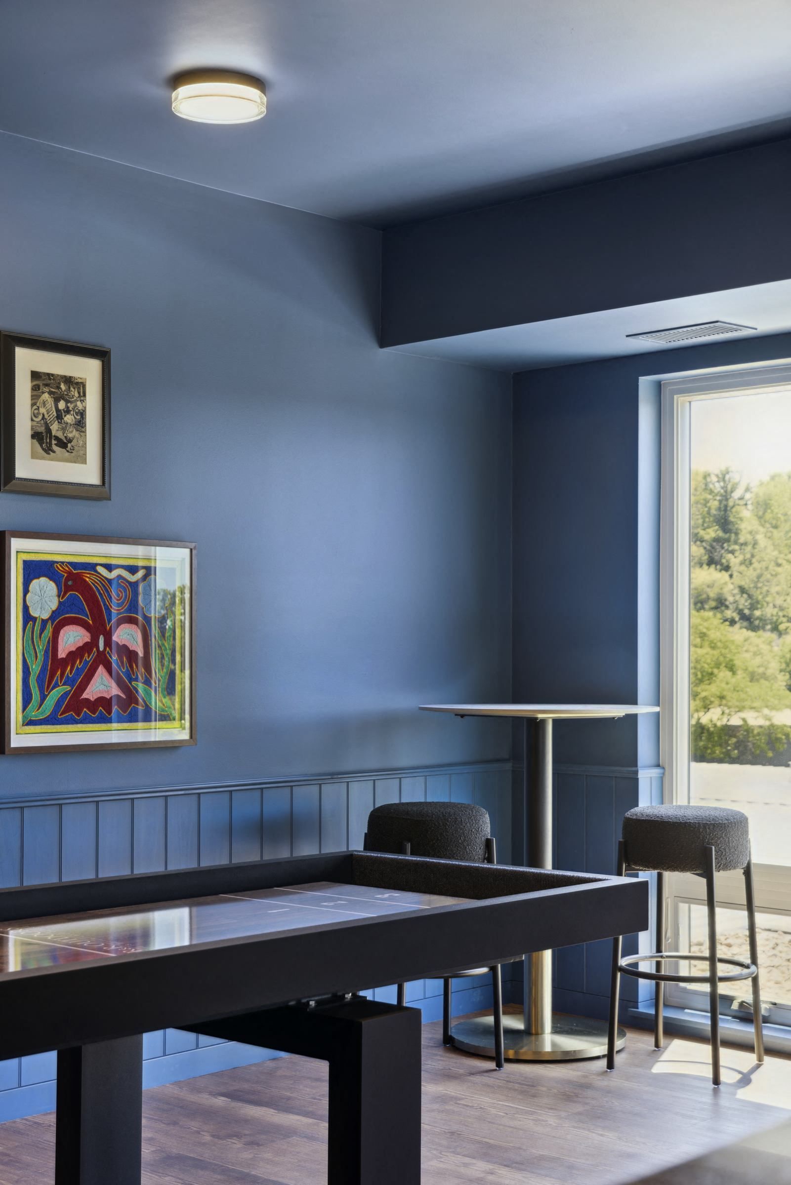 Blue-walled lounge area with a high-top table, two stools, and a large window at Esox House in St. Paul, MN.