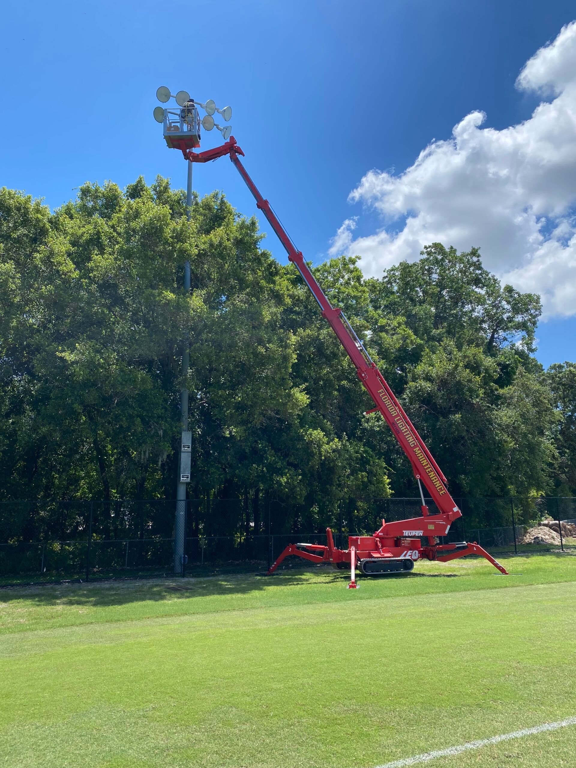 Red lift truck servicing sports field lights near a tree line on a green field under a blue sky.