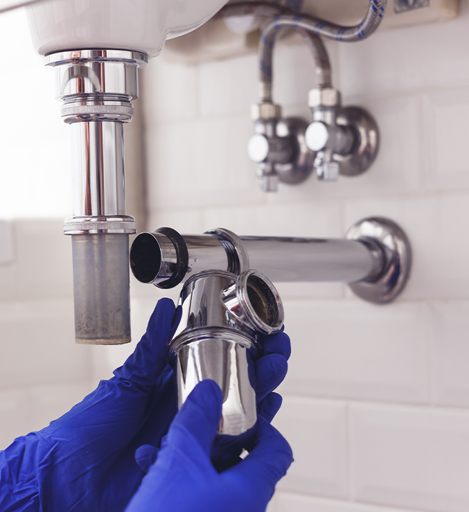 Hands in blue gloves assembling plumbing under a sink.