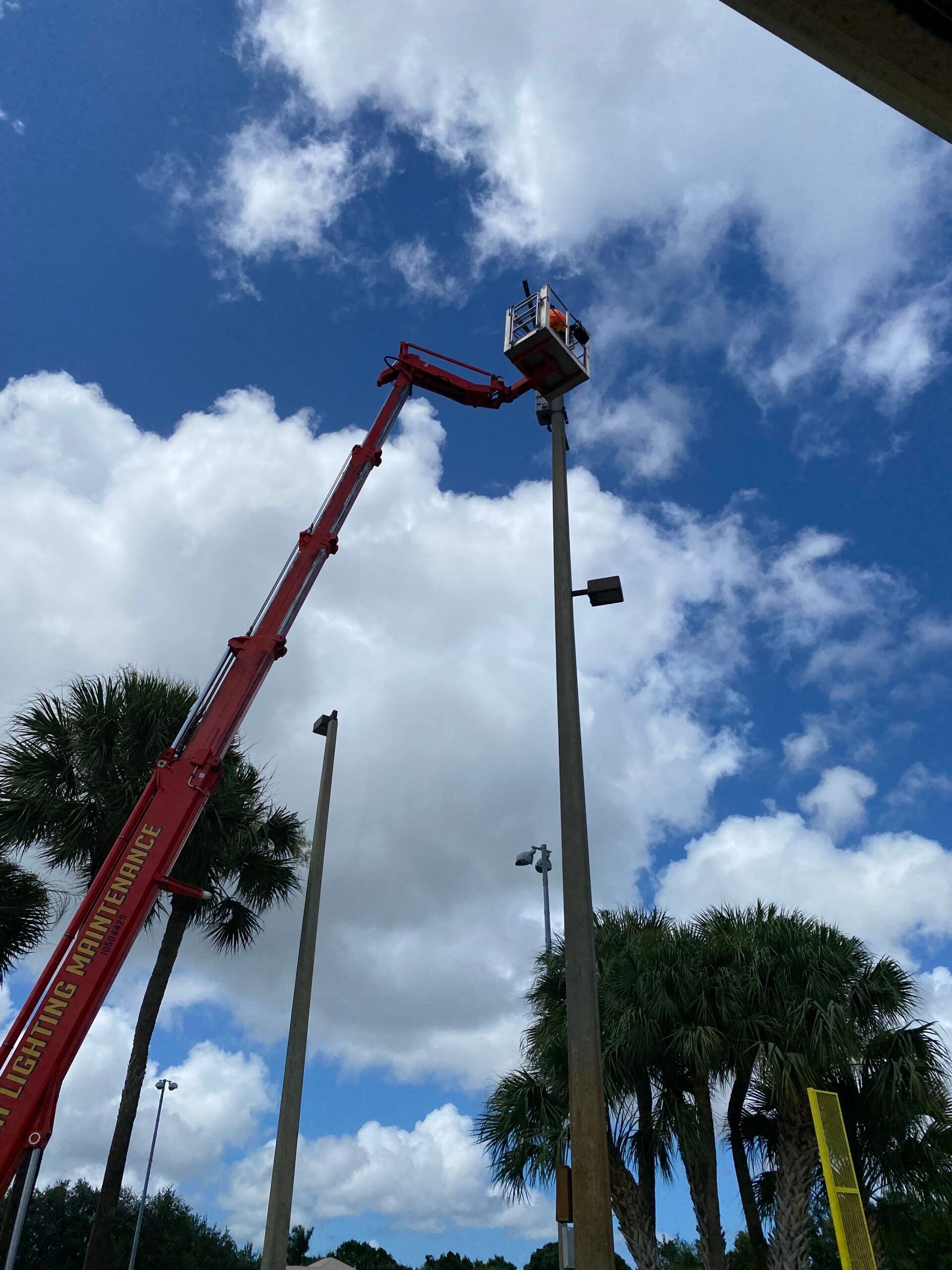 A worker in a lift basket is working on a tall light pole against a blue cloudy sky.