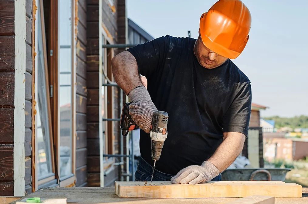 Construction worker in orange hard hat drills wood on a sunny day.