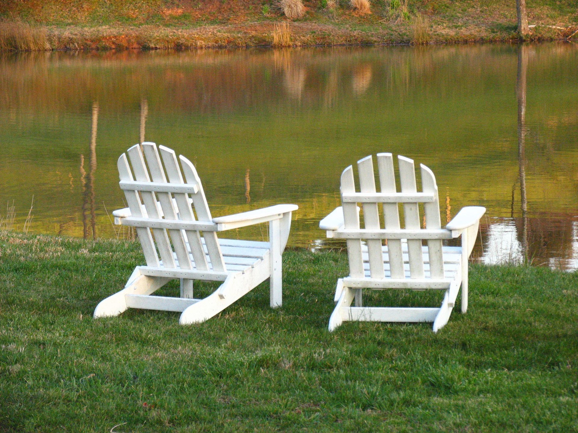 Two white adirondack chairs are sitting in the grass near a lake.