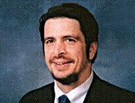 A person with a beard and dark hair wearing a dark suit and tie, smiling against a blue textured background.