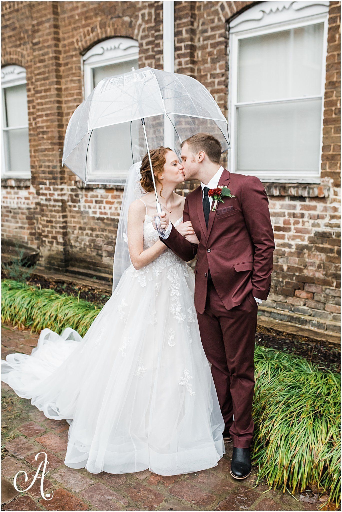 A bride and groom are kissing under an umbrella in front of a brick building.