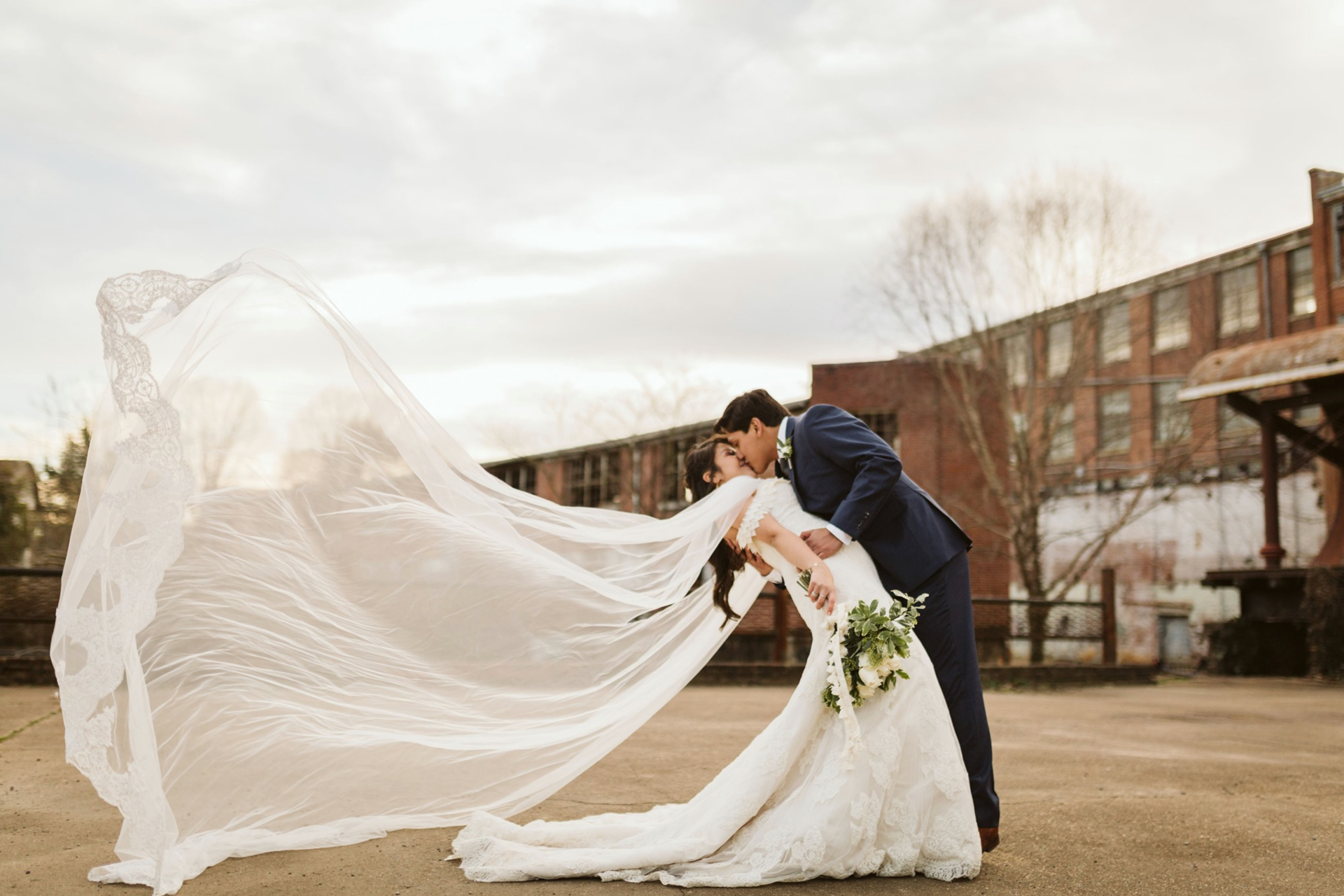 A bride and groom kissing with a long veil blowing in the wind.