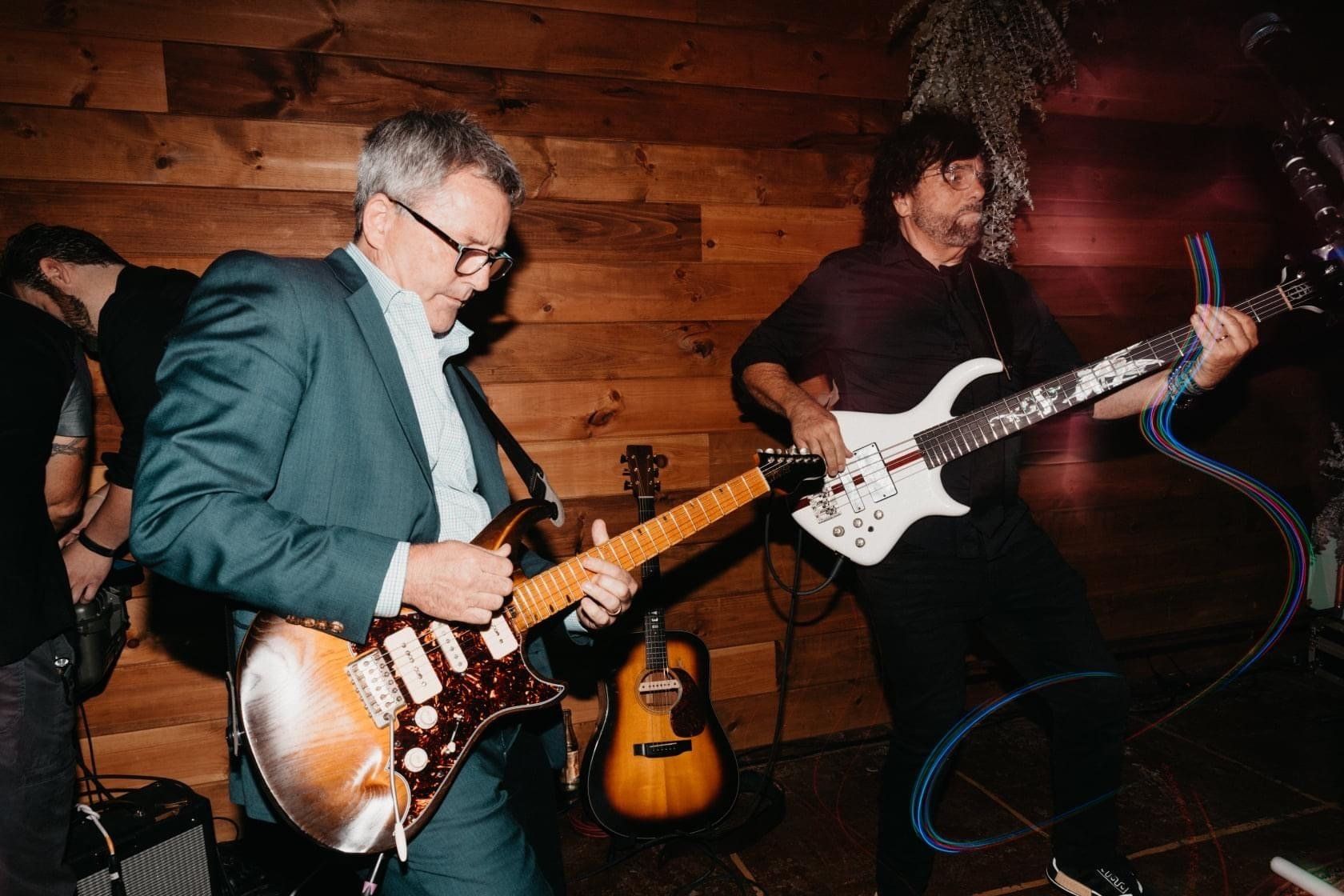 Two men are playing guitars on stage in front of a wooden wall.