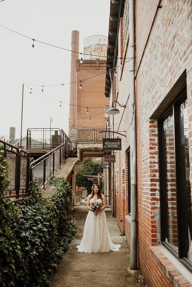 A bride in a wedding dress is standing in an alleyway between two brick buildings.