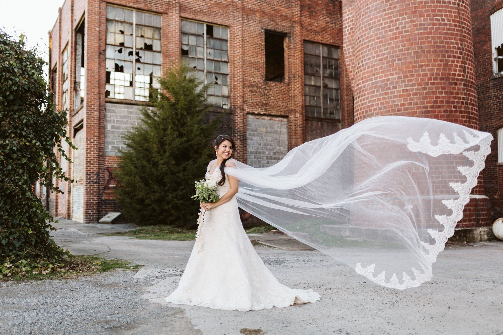 A bride in a wedding dress is standing in front of a brick building with her veil blowing in the wind.