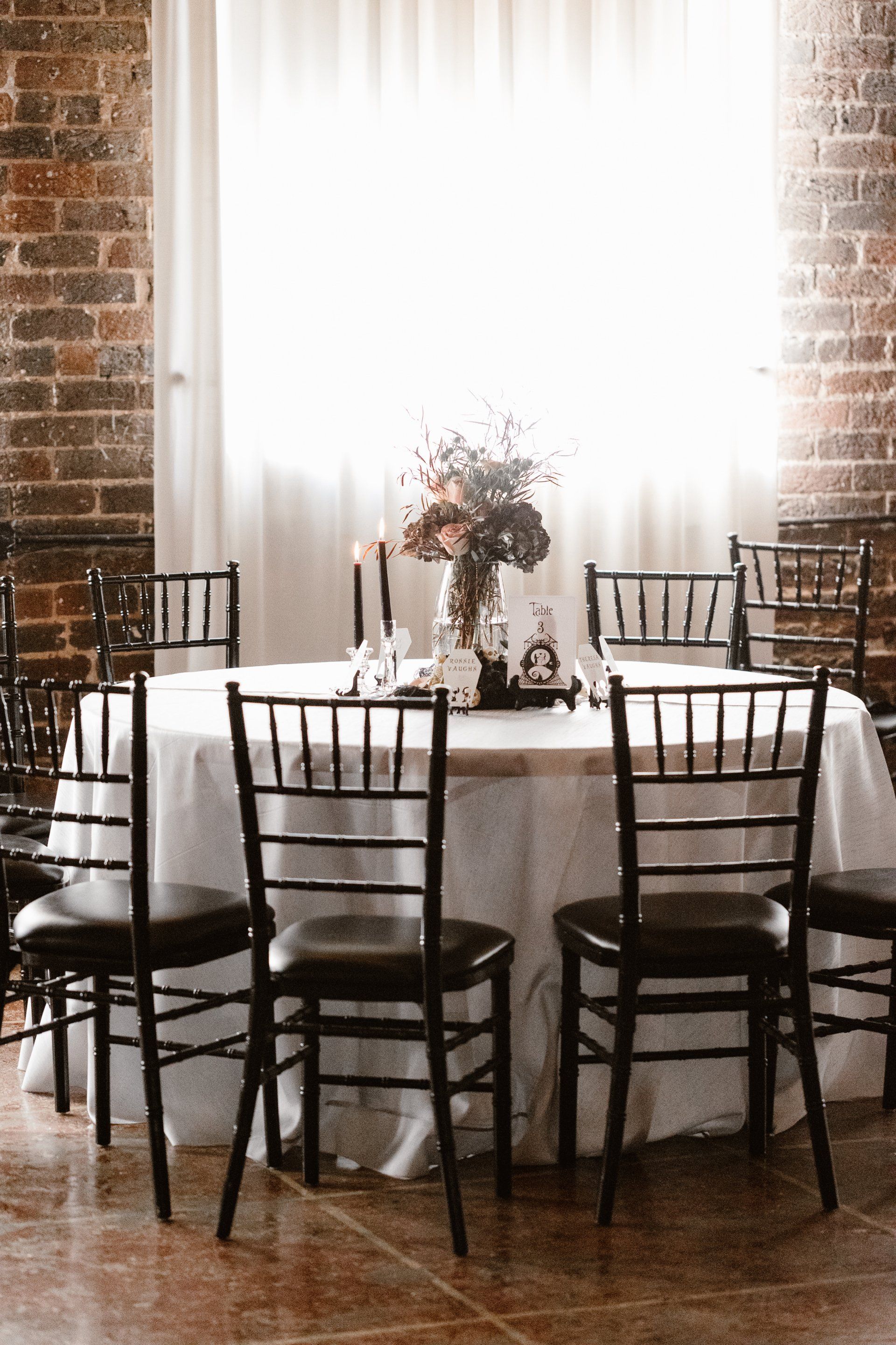 A table with a white tablecloth and black chairs in a room with a brick wall.