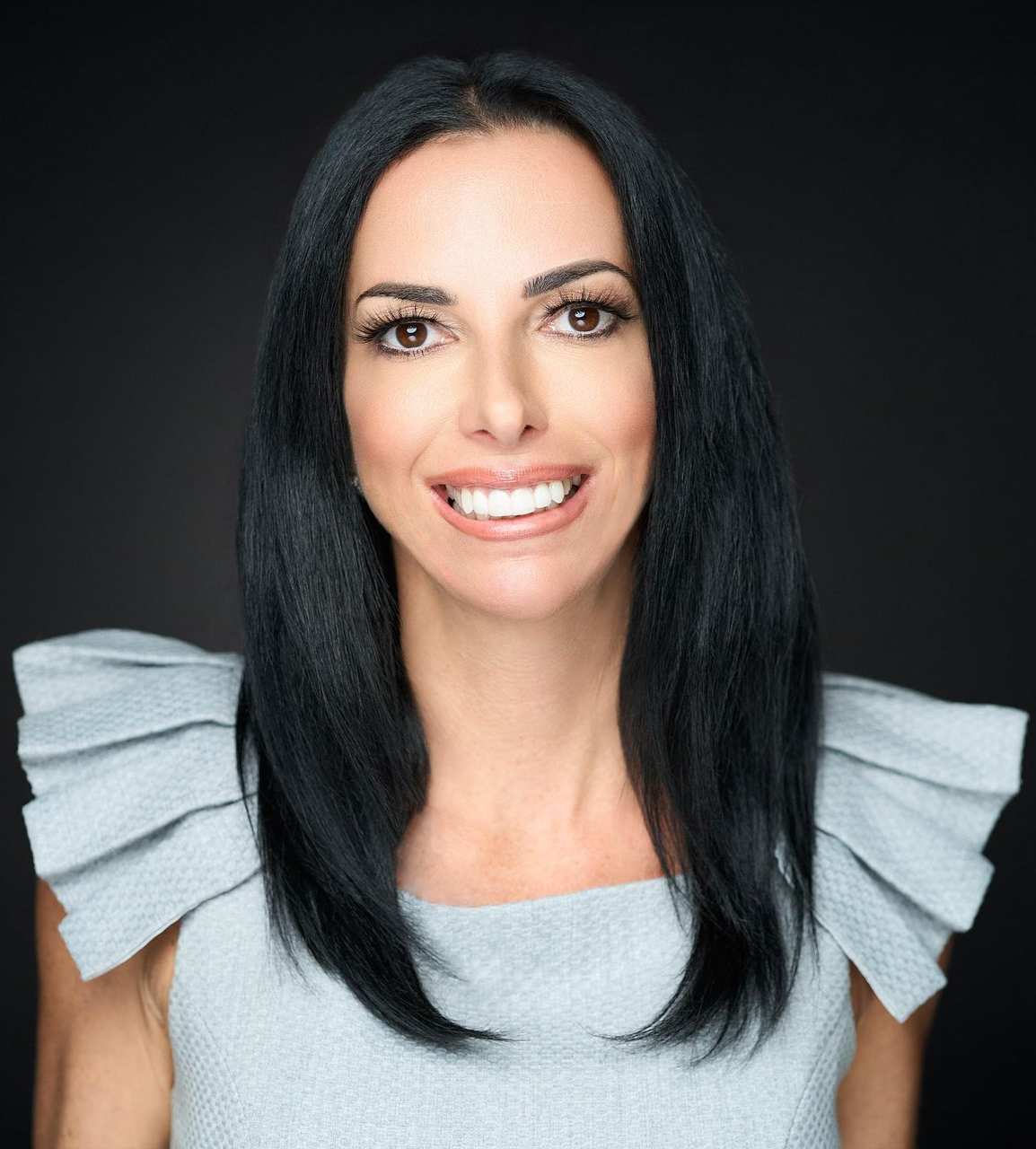 Woman with dark hair smiles, wearing a light blue textured dress against a dark background.