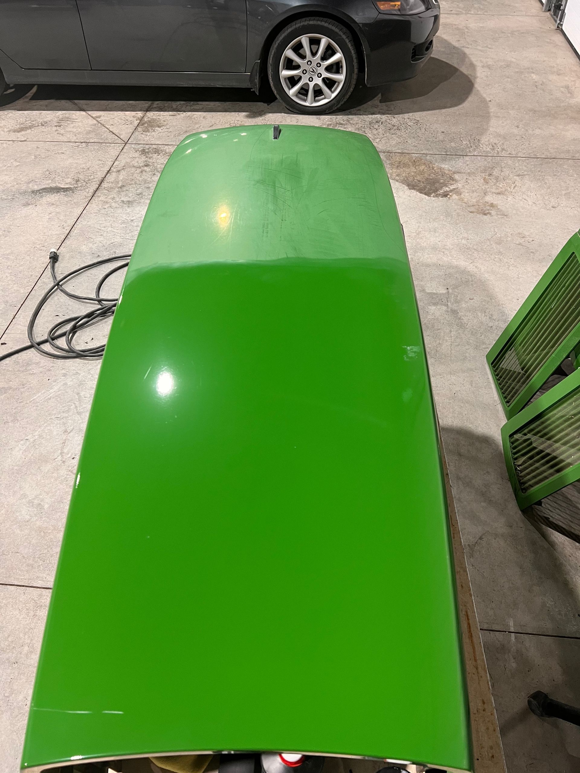 A green hood of a tractor is sitting in a garage next to a car.