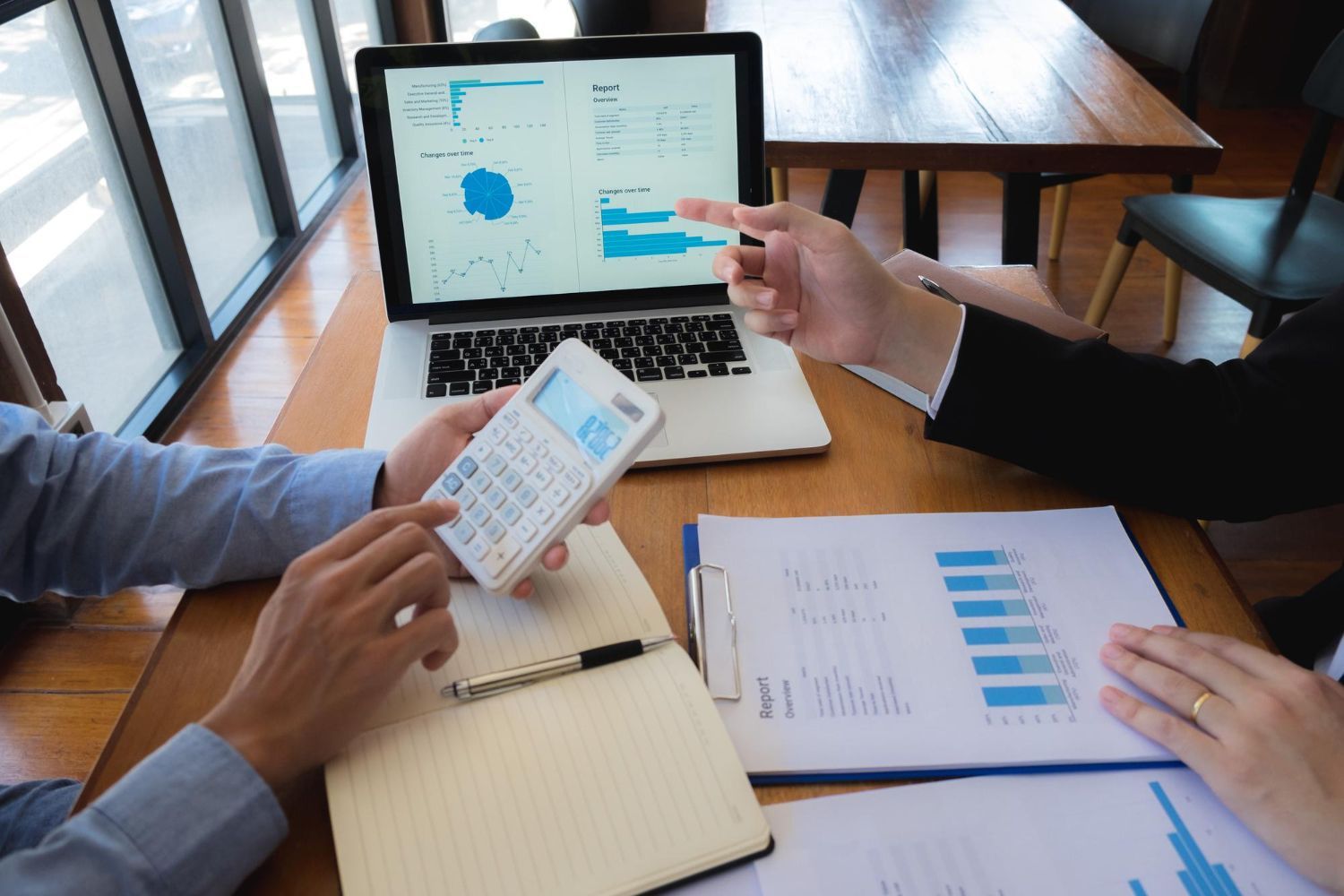 Two people reviewing financial data on a laptop and documents, using a calculator, in an office.