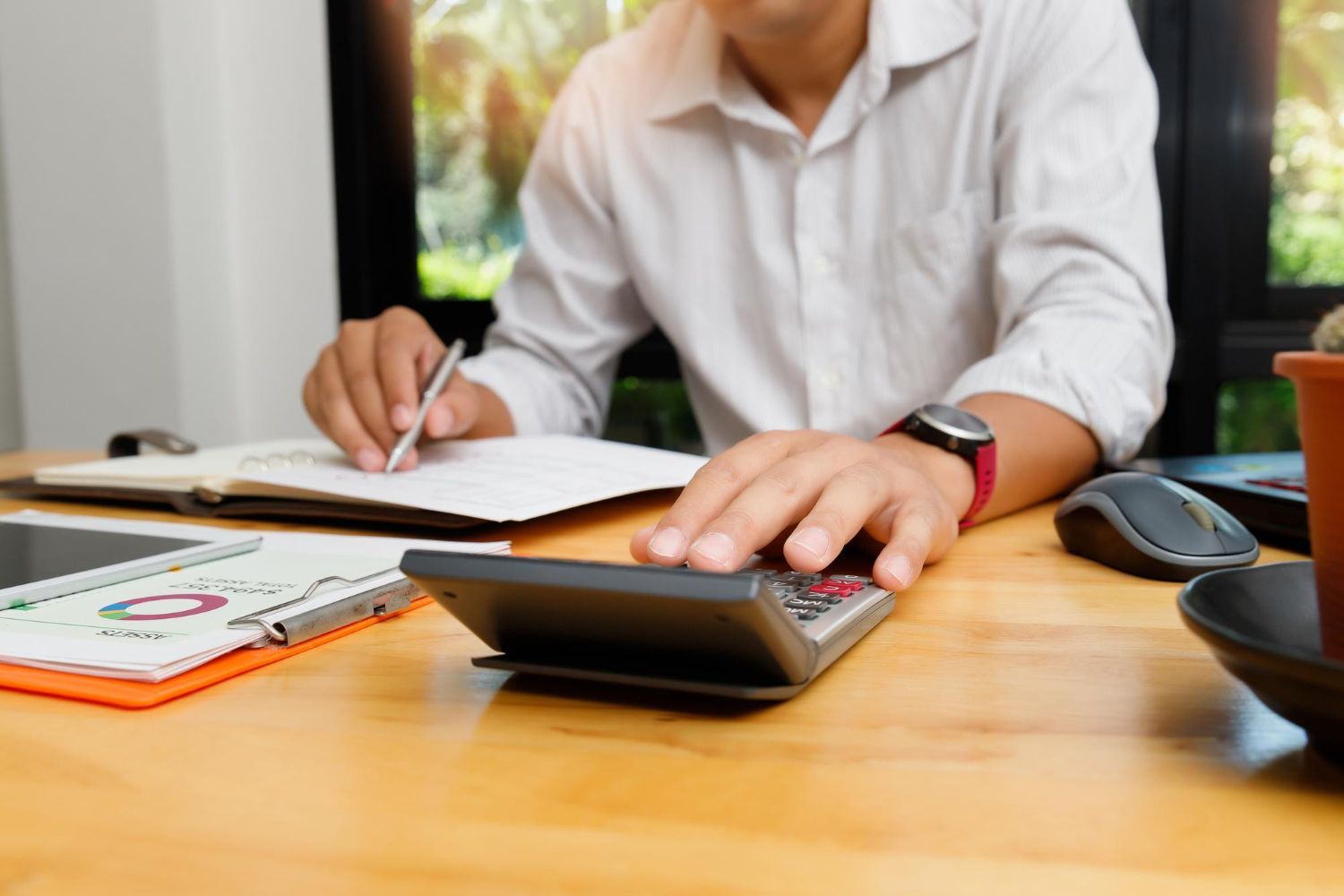Person in a white shirt using a calculator at a wooden desk, writing and reviewing financial data.