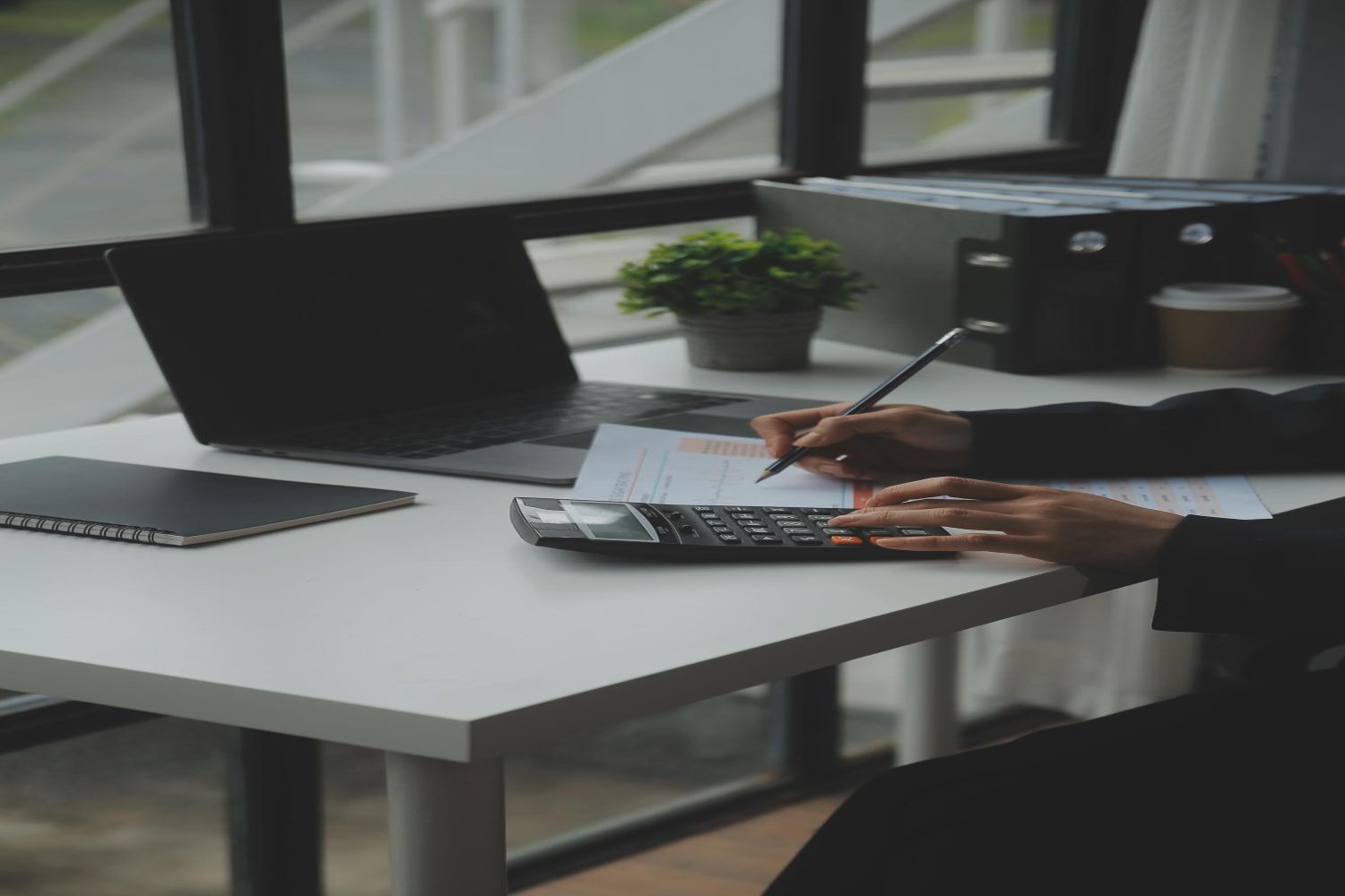 Person working at a desk, using a calculator and pen, near a laptop and printer.