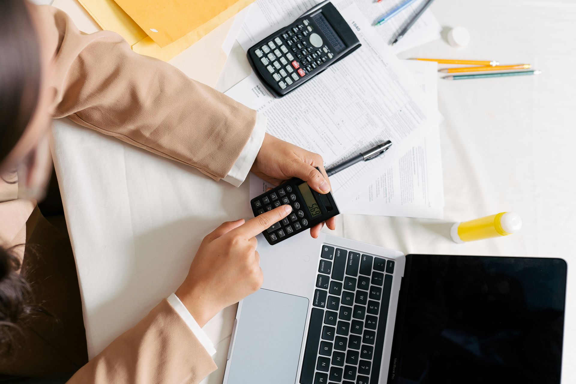 Person using a small calculator on a white desk with a laptop, large calculator, and papers.