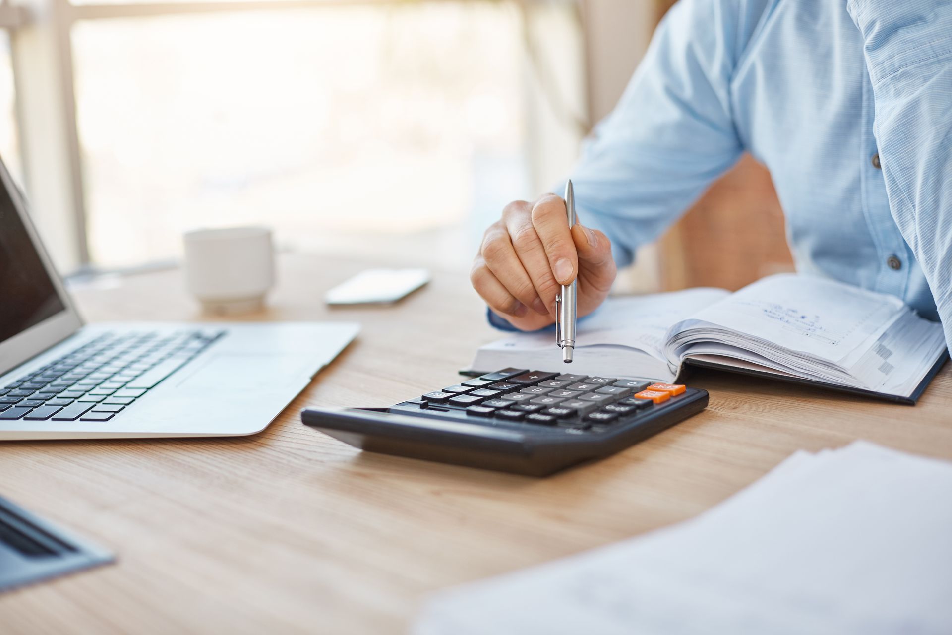 Person in a white shirt using a calculator at a wooden desk, writing and reviewing financial data.