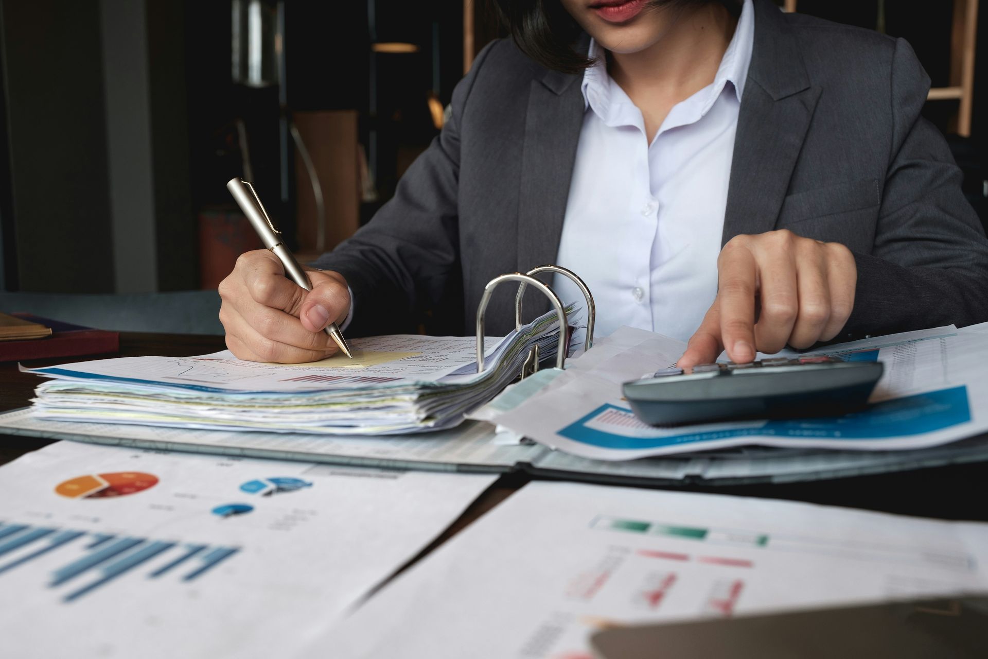 Person in a gray suit working with papers, binder, pen, and calculator at a desk.