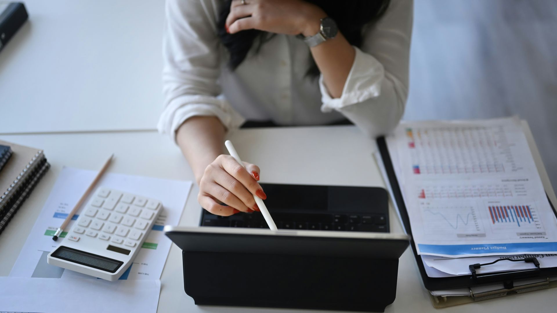 Person in a white shirt using a calculator at a wooden desk, writing and reviewing financial data.