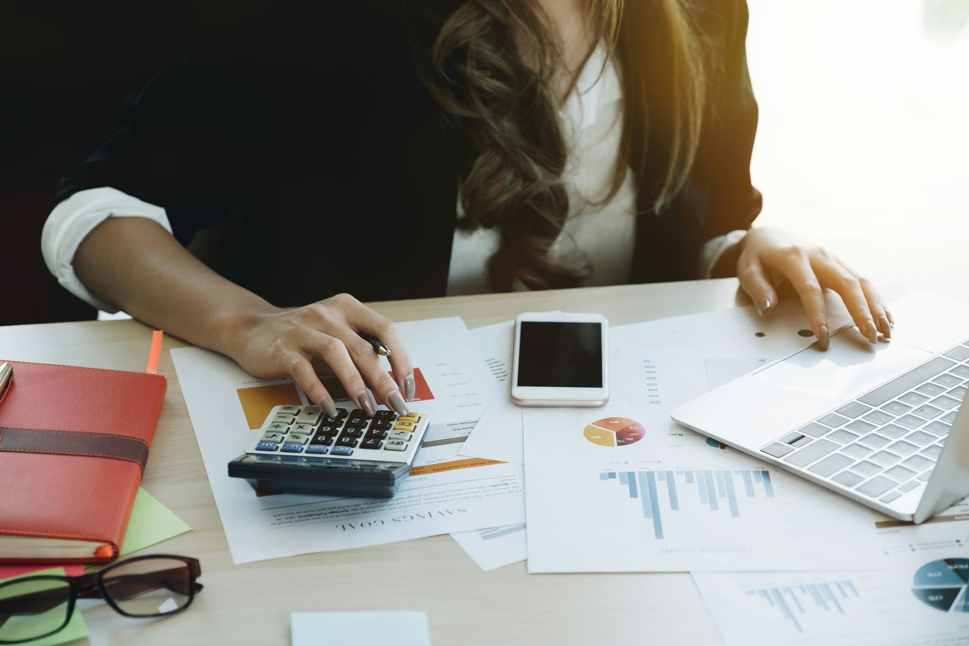 Person in a white shirt using a calculator at a wooden desk, writing and reviewing financial data.