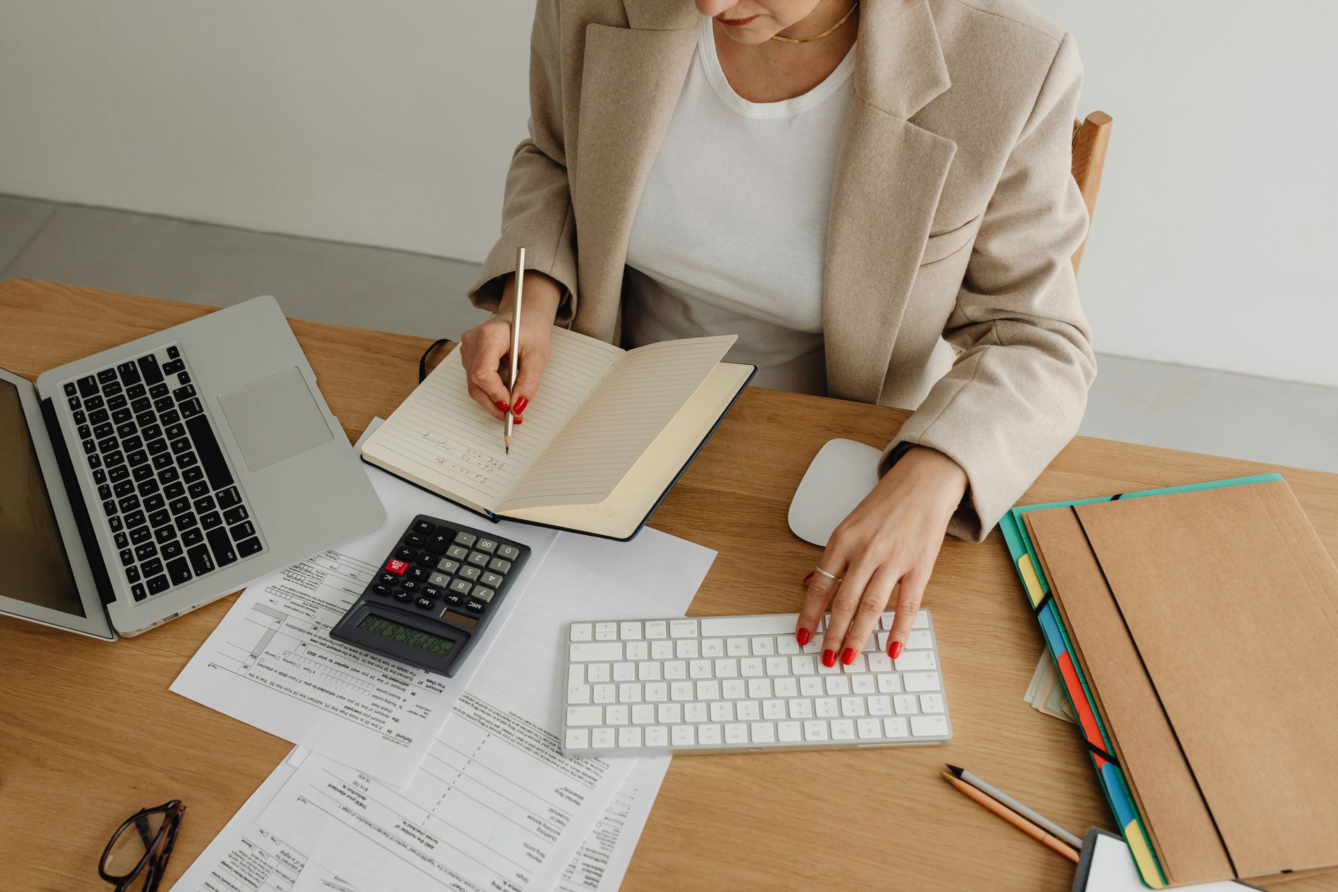 Person in a white shirt using a calculator at a wooden desk, writing and reviewing financial data.