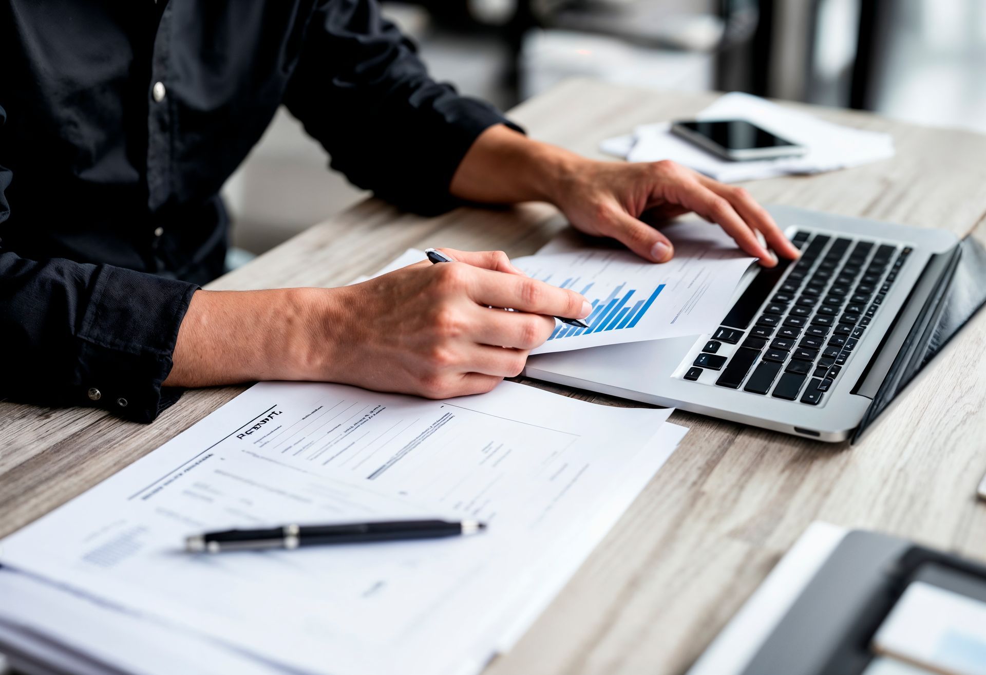Person in black shirt analyzes charts on paper and laptop at a desk.