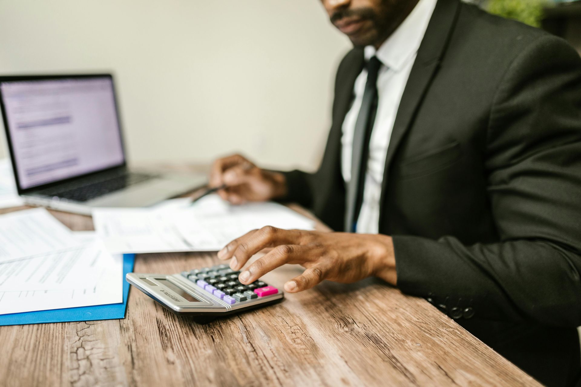 Person in a white shirt using a calculator at a wooden desk, writing and reviewing financial data.