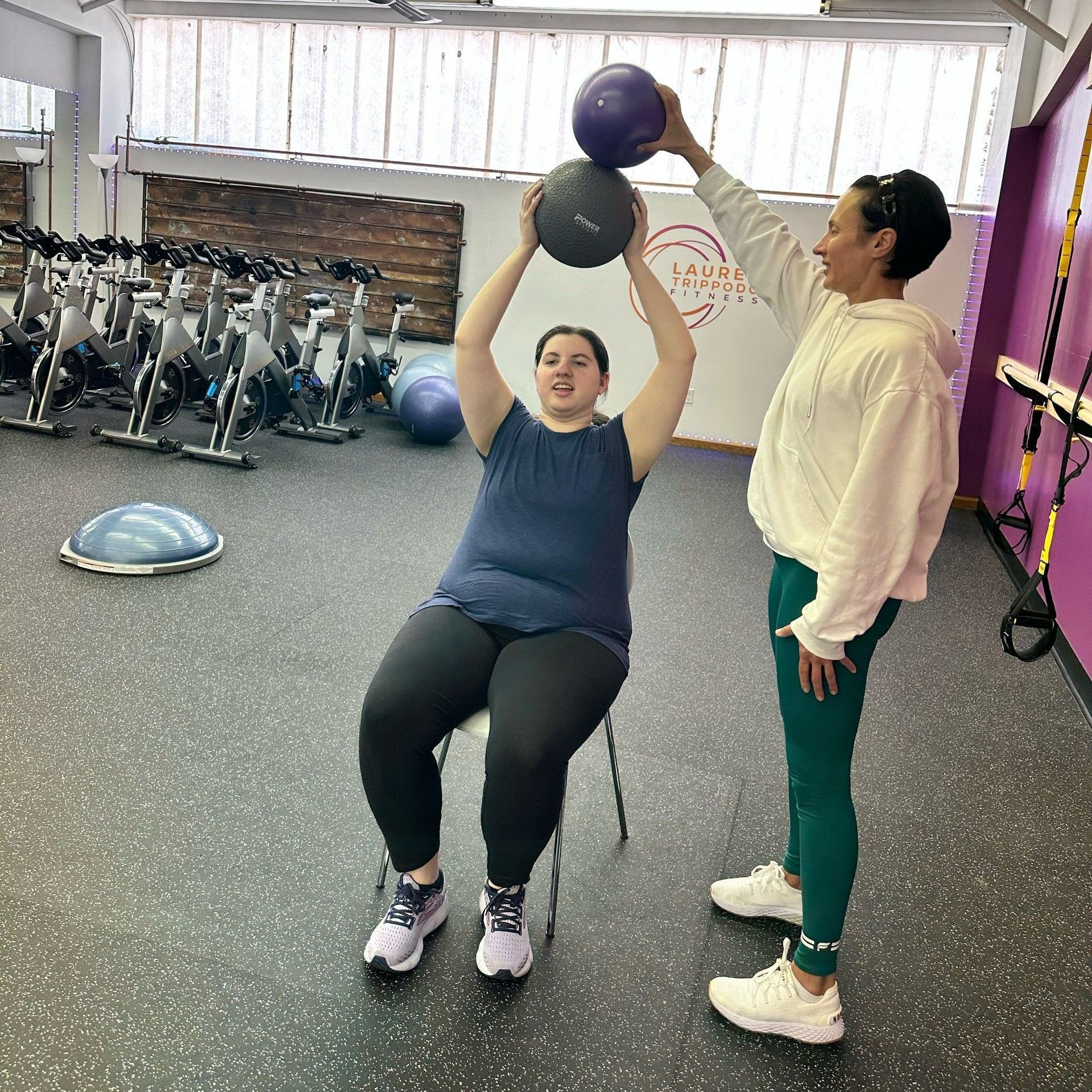 A woman is sitting on a chair in a gym while another woman holds a ball over her head.