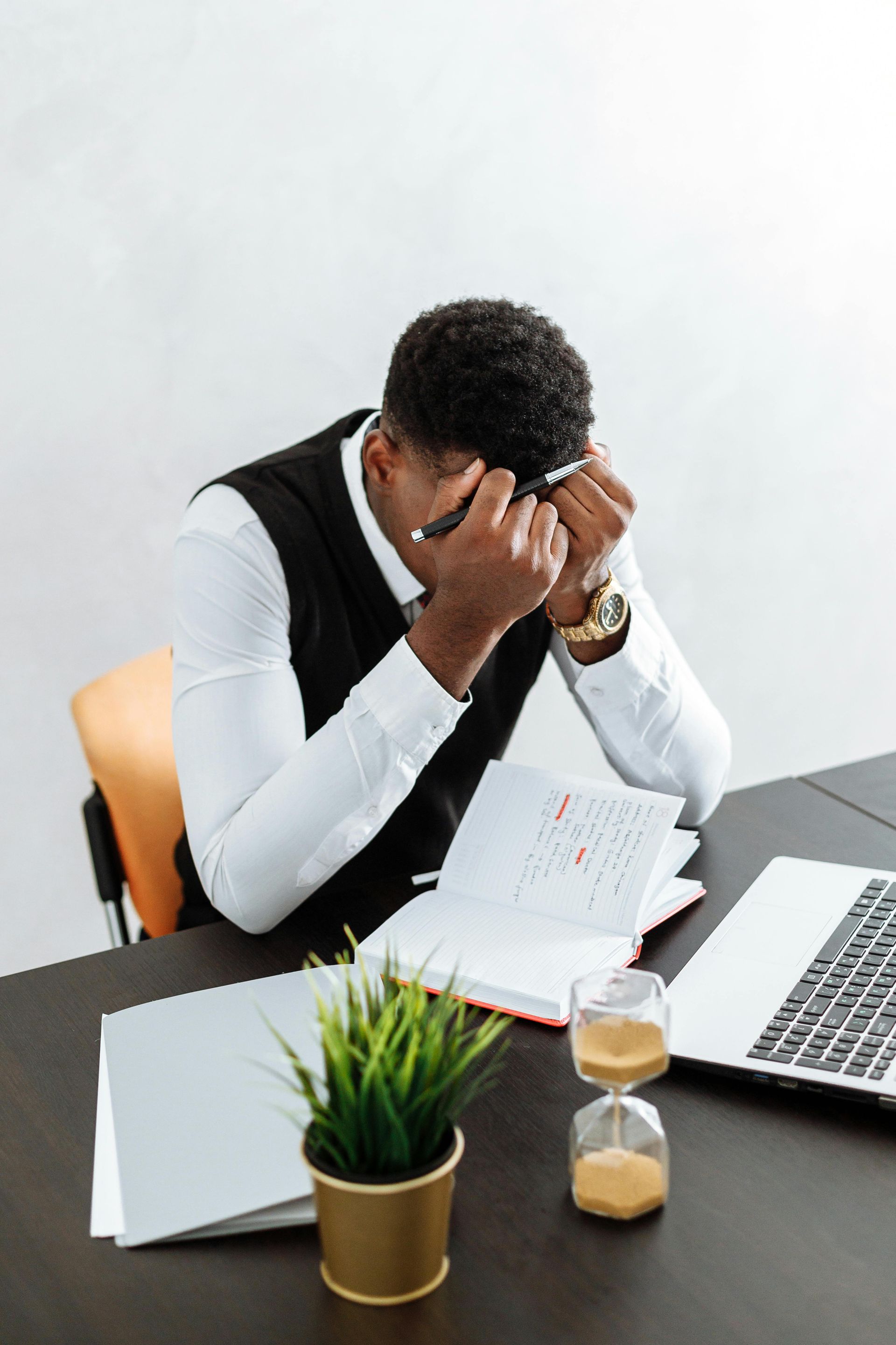 Man in vest at desk, stressed, head in hands, papers, hourglass, laptop.