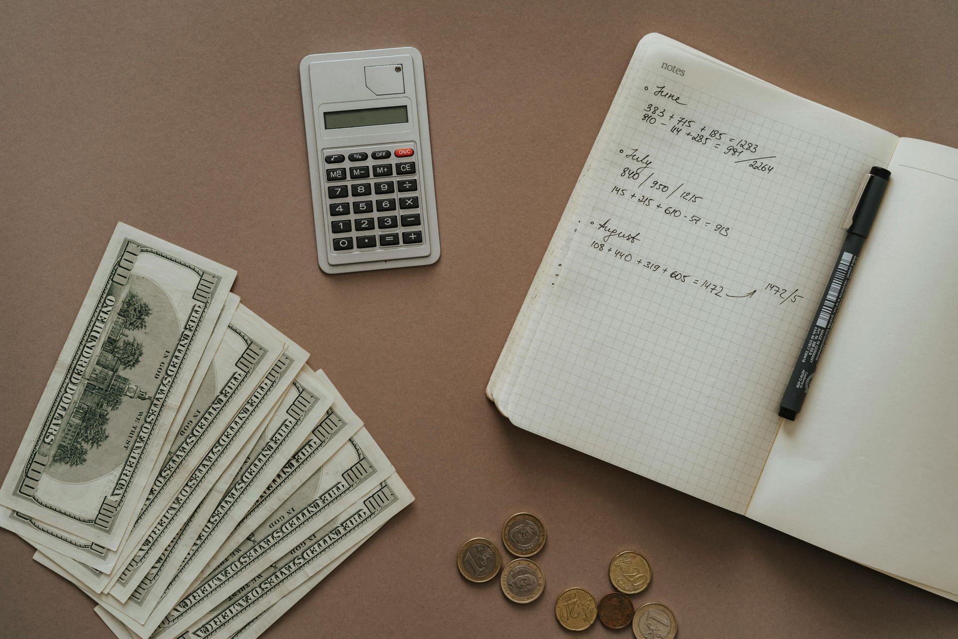 Cash, calculator, notepad with writing, and coins arranged on a brown surface.