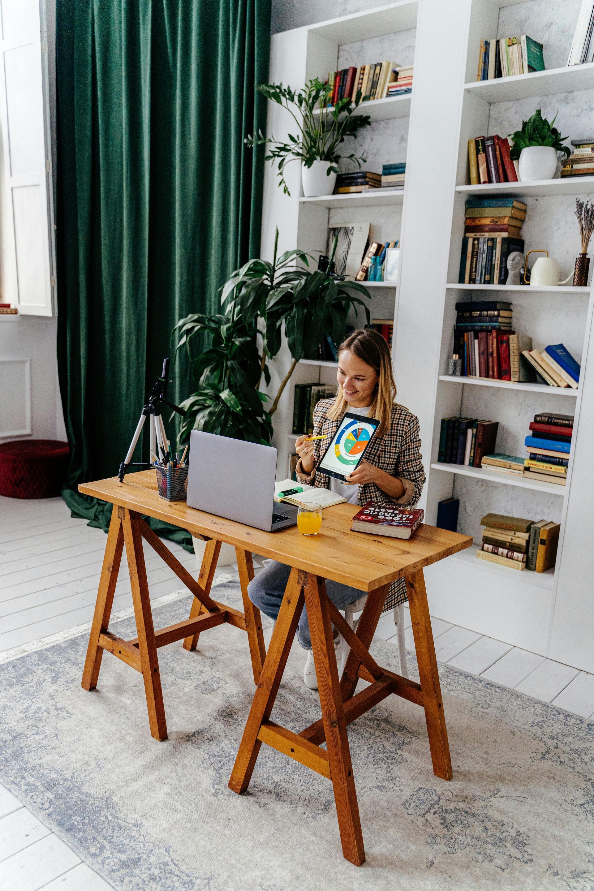 Woman at wooden desk, using tablet and laptop. Bookshelves, green curtain, and rug in a bright room.