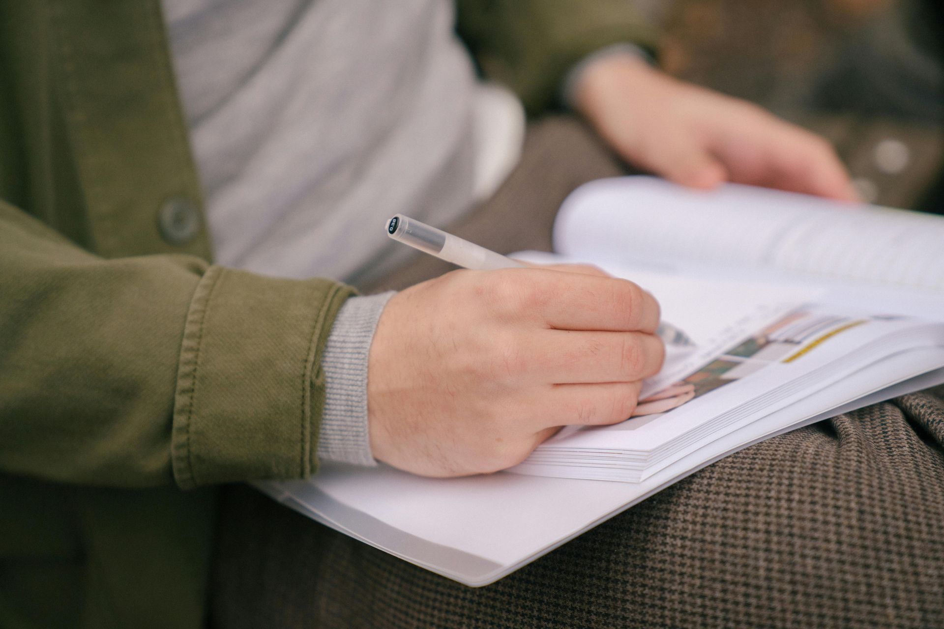 Person writing with a pen in a notebook. They are wearing a green jacket.