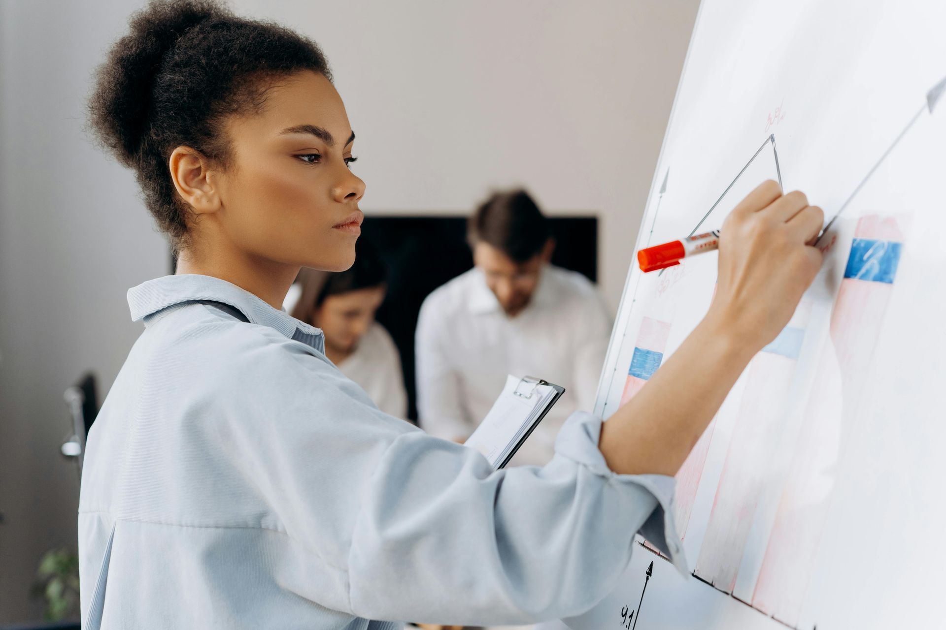 Woman drawing on a whiteboard with a red marker, two people in the background, light-colored office.