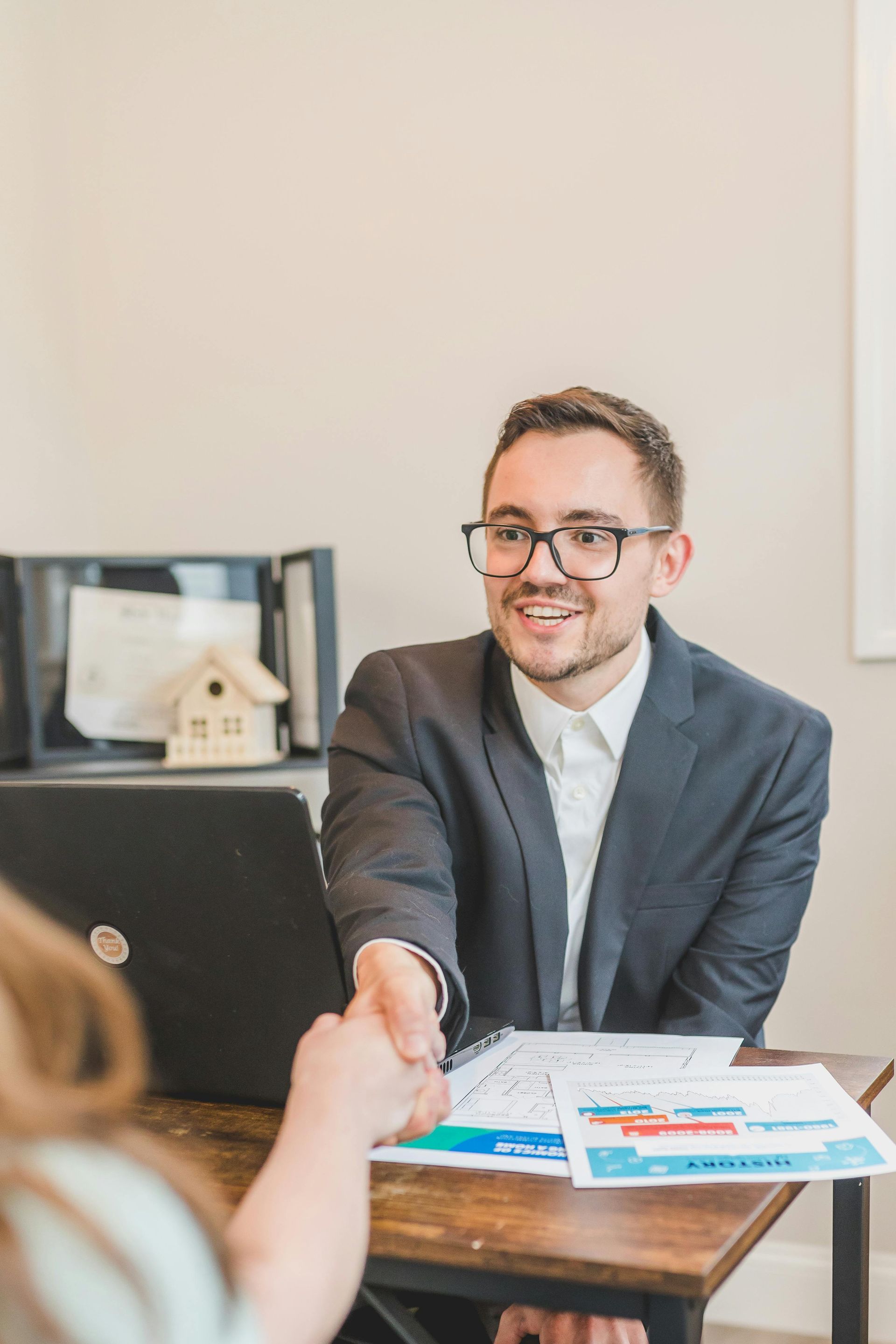 Man in suit shaking hands with a person at a desk with laptop, documents, and house model.