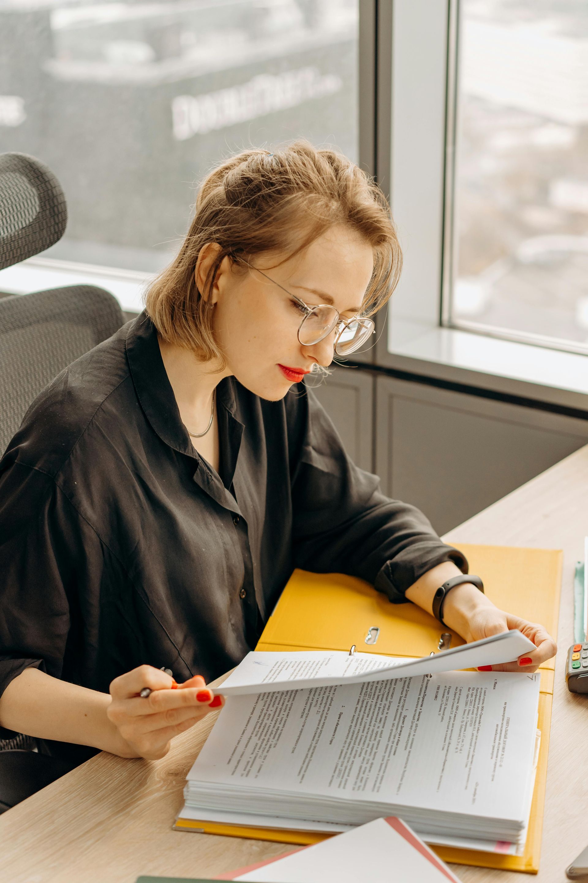 Woman wearing glasses examines documents at a desk, red lipstick, yellow folder, office setting.