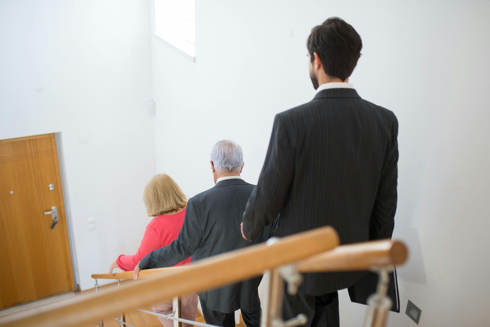 People walking up stairs. One person in a dark suit leads, followed by an older couple holding the railing.