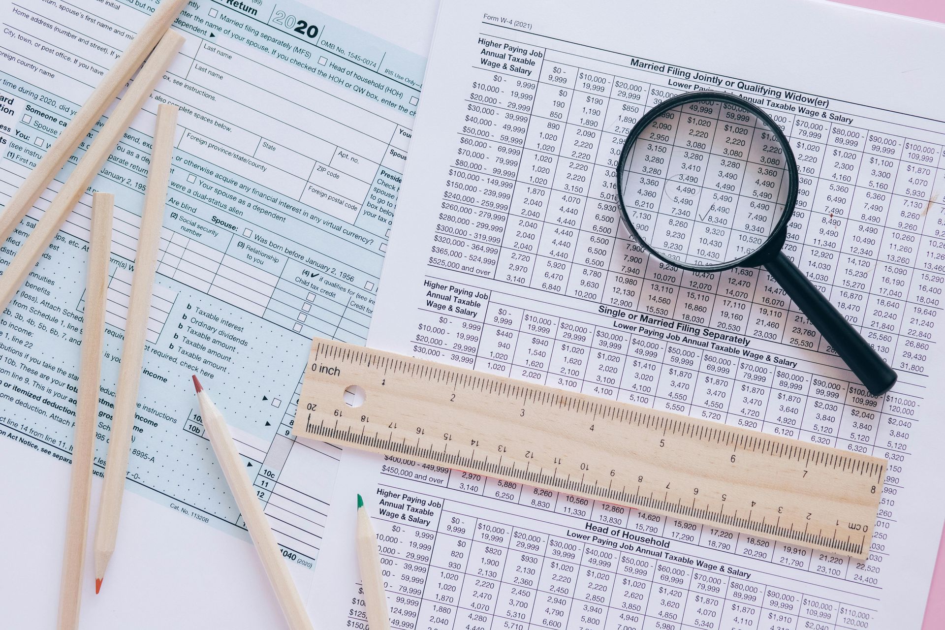 Papers, magnifying glass, ruler, and pencils on a light pink surface, suggesting document review.