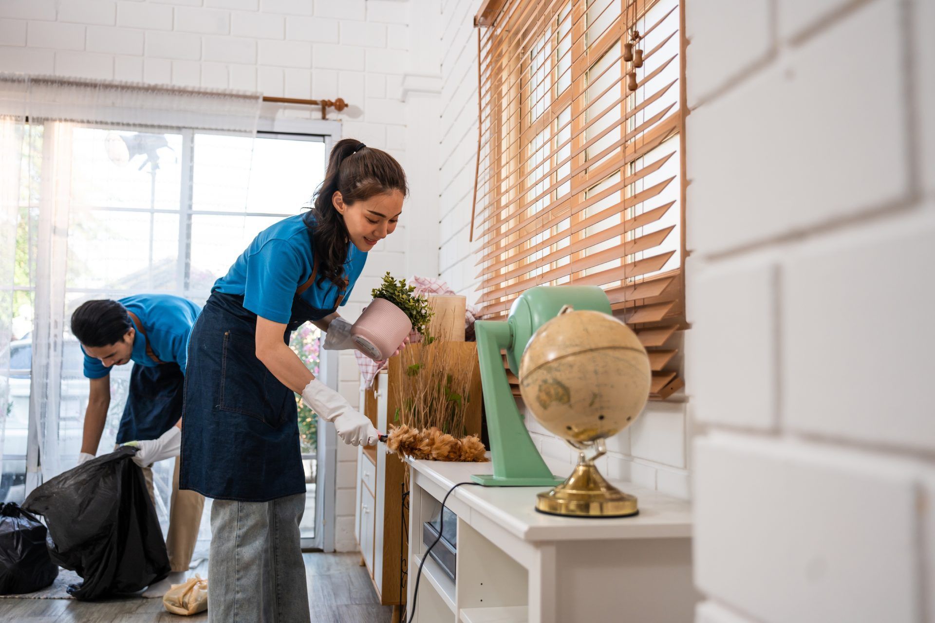 Two people cleaning a room: one with a plant, the other with a trash bag. Brightly lit interior.