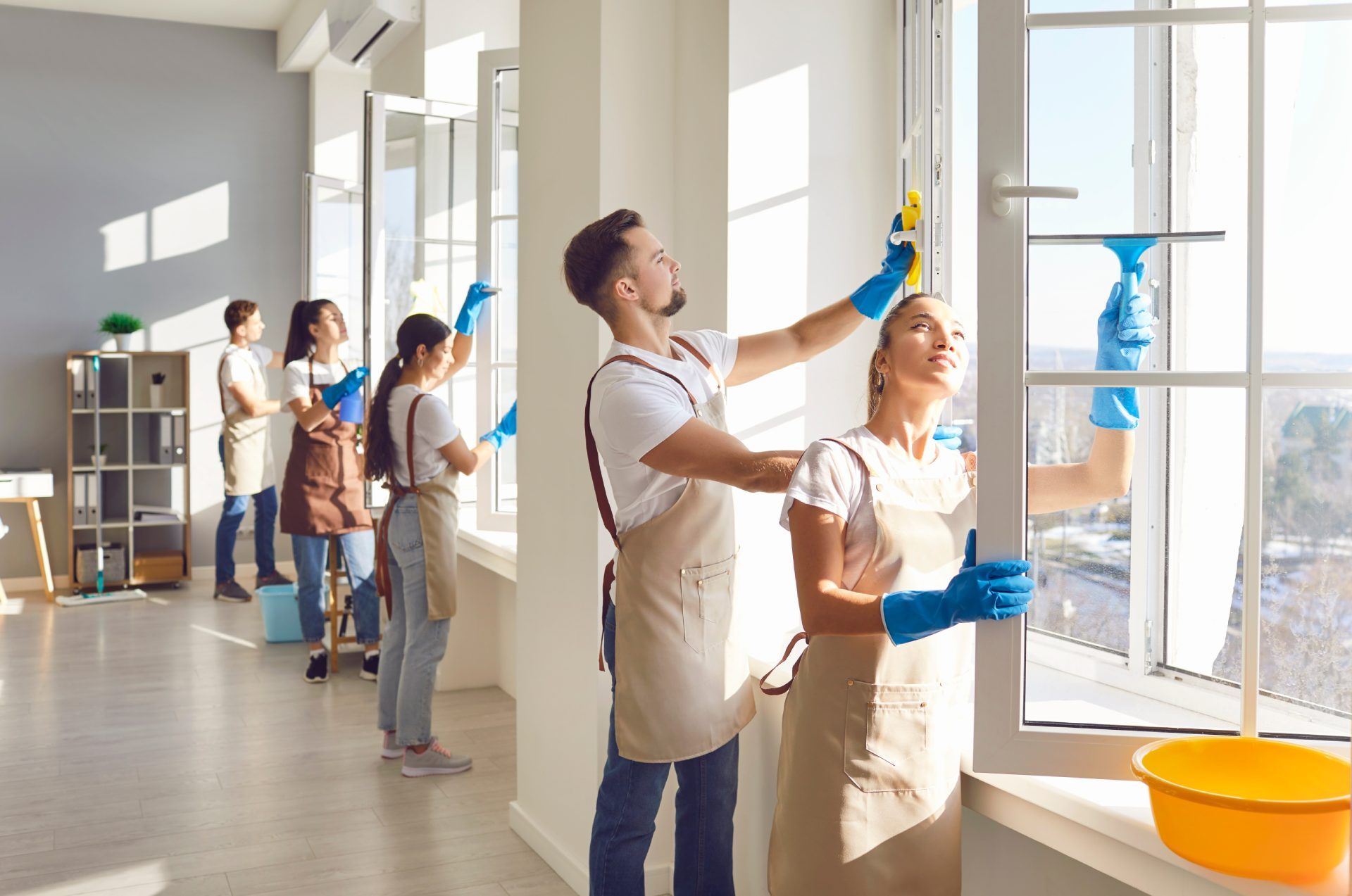 People cleaning windows in a well-lit room, using squeegees, buckets, and wearing aprons.