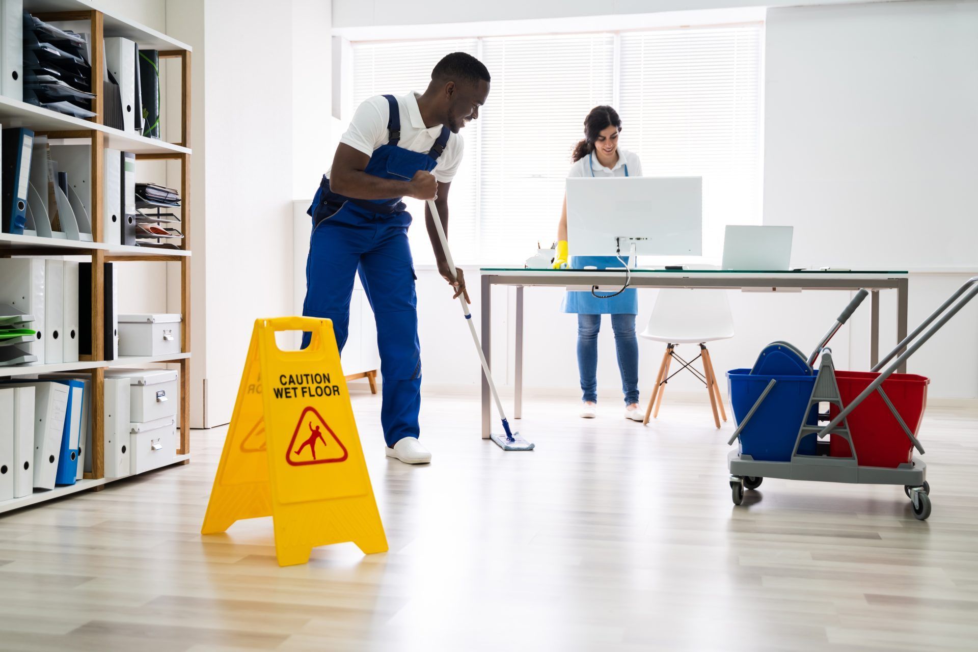 Office cleaning: Person mopping floor, caution sign, another person at desk, cleaning cart.