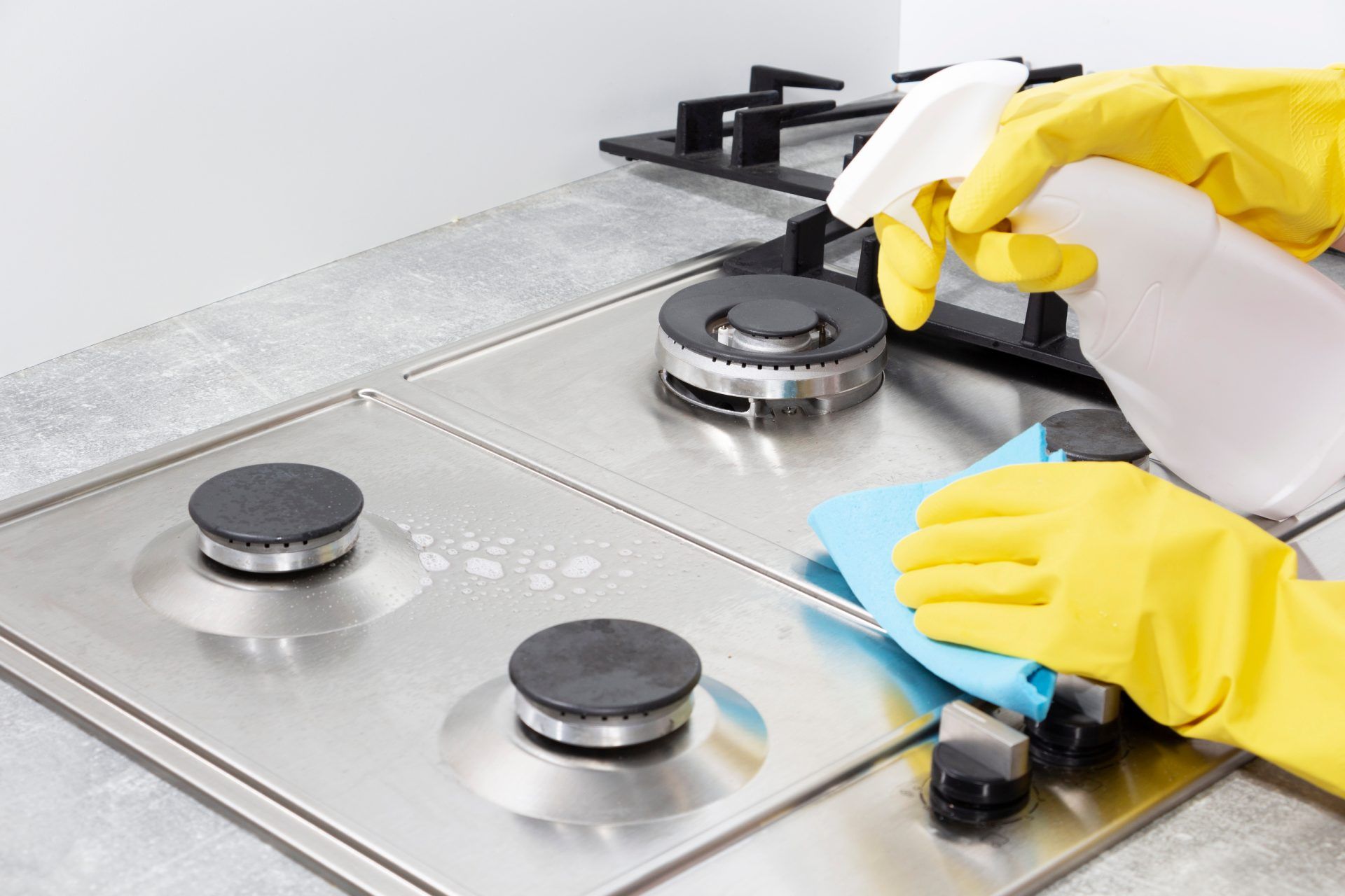 Hands in yellow gloves cleaning a stainless steel stovetop with spray bottle and blue cloth.